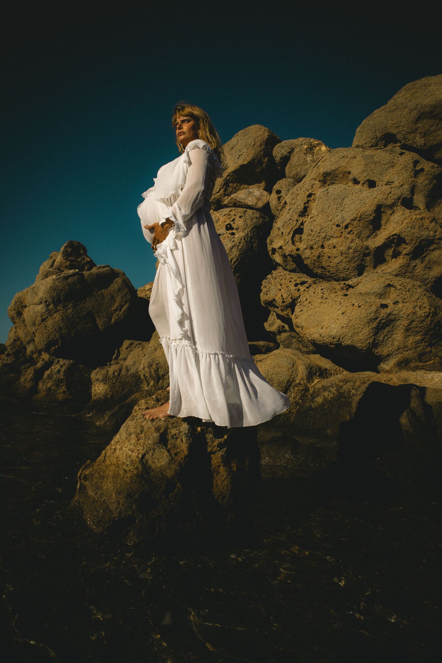 A figure in flowing white clothing poses dramatically against rocky coastal formations at dusk.