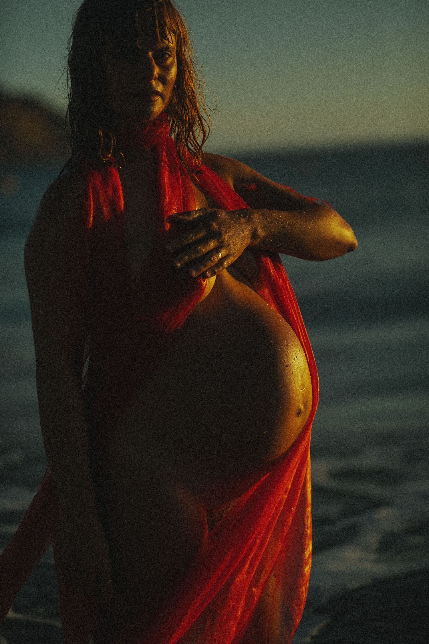 Figure in flowing red fabric poses dramatically at sunset by the ocean.