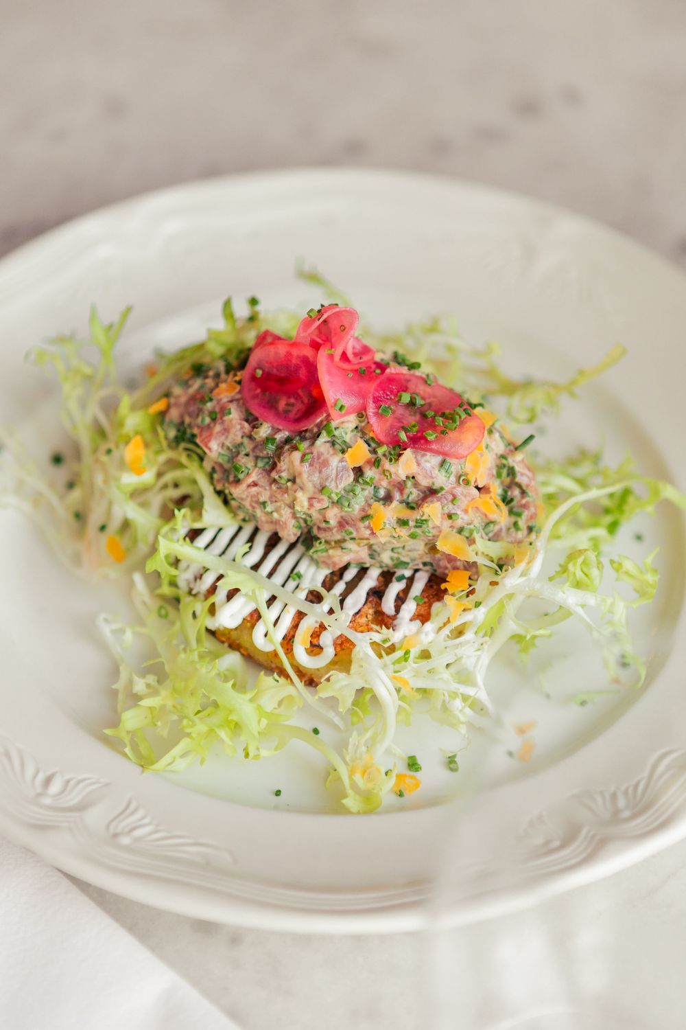 Artfully composed salad with frisee lettuce, pink garnish and creamy dressing on an ornate white china plate.