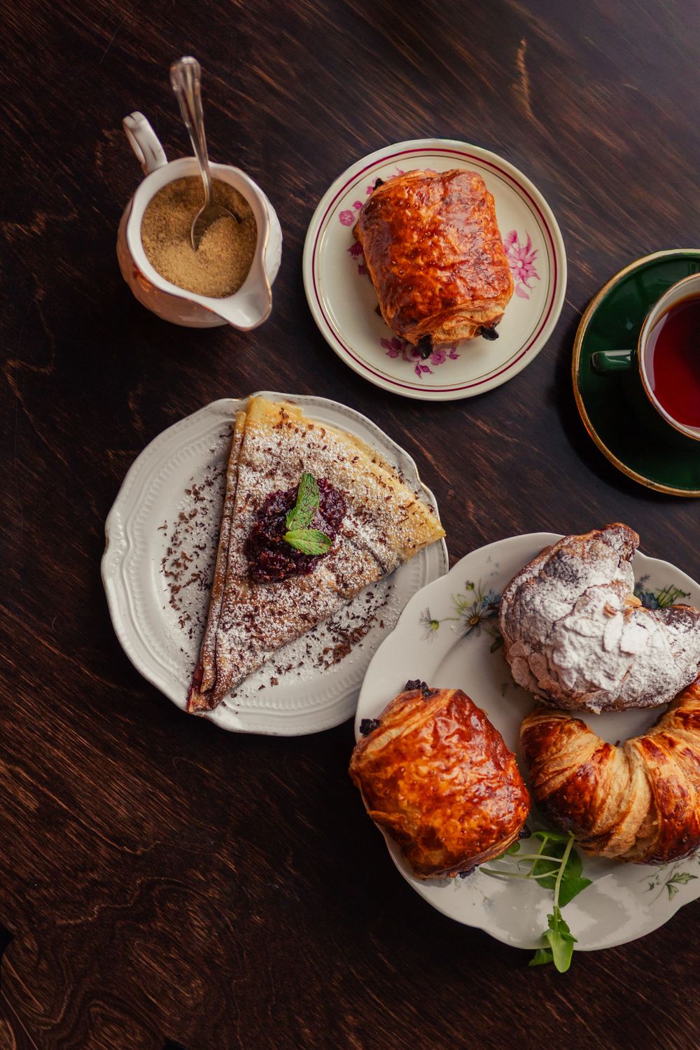 Freshly baked pastries and desserts including chocolate cake and croissants arranged on white plates against a dark wooden surface.
