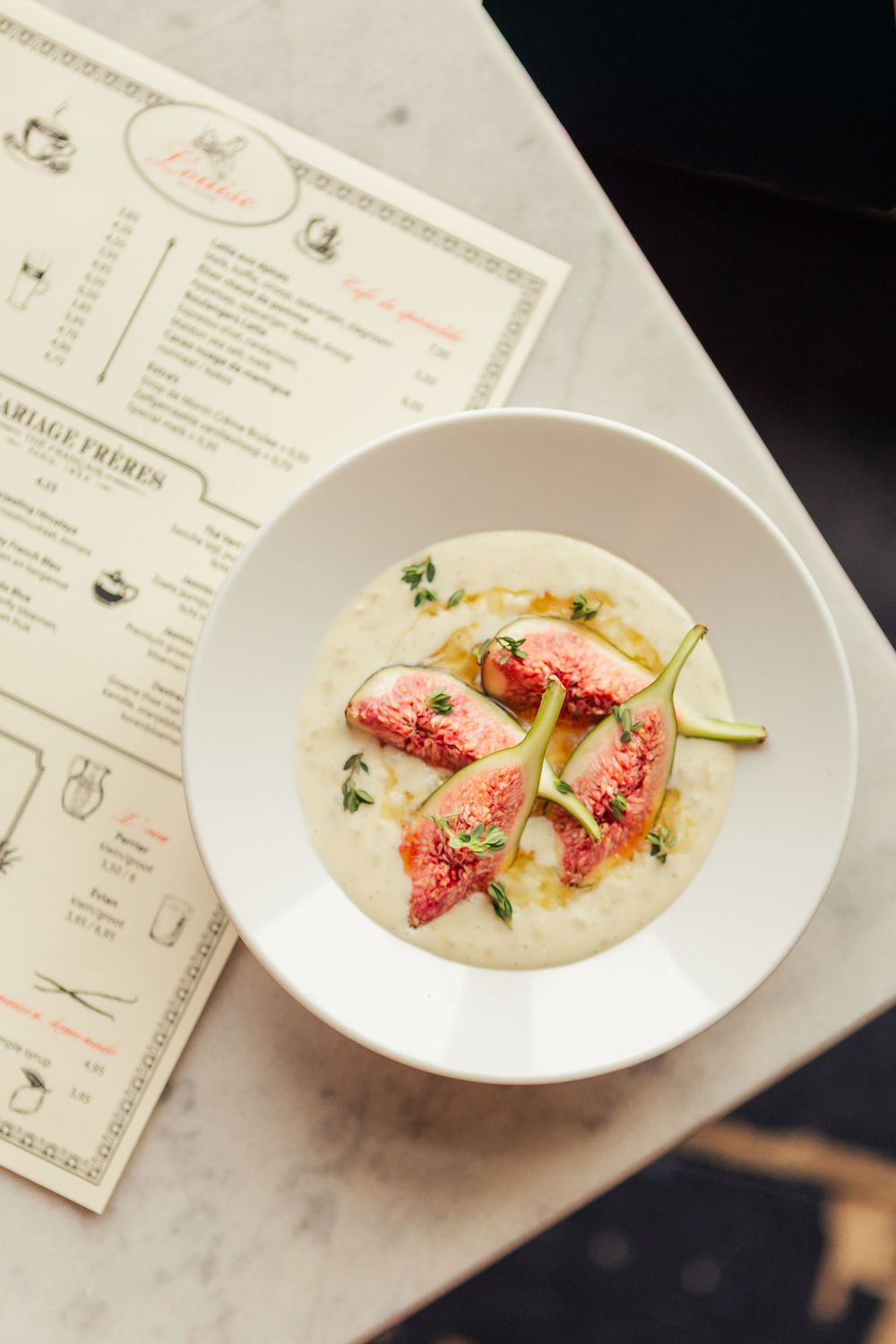 White ceramic bowls of creamy soup garnished with fresh figs and herbs on a marble surface with menu cards.