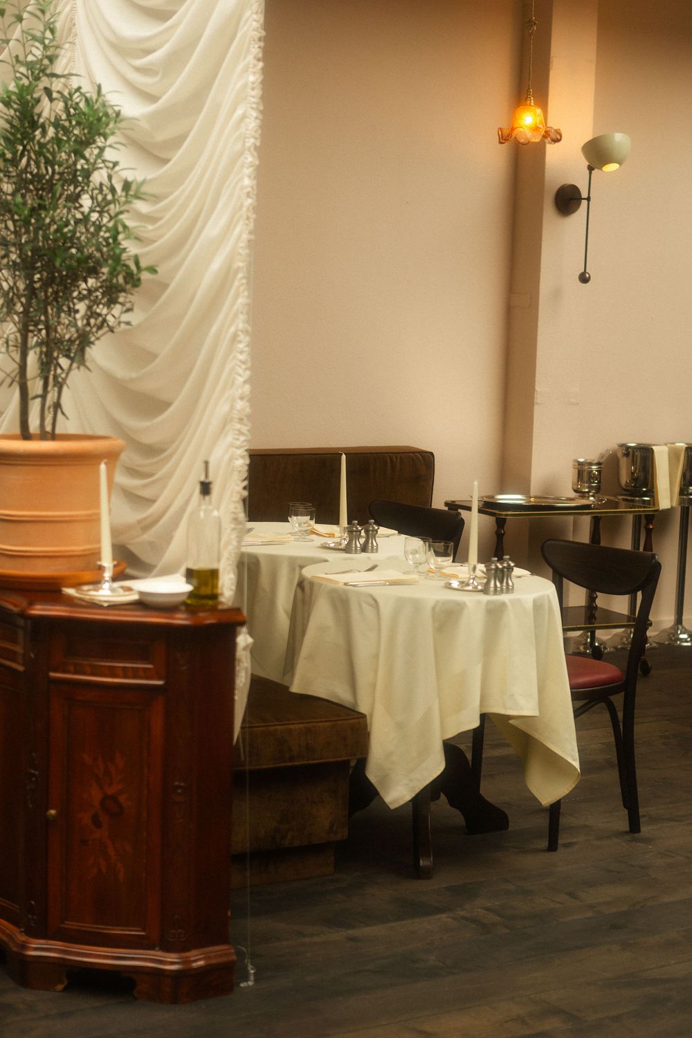 Elegant restaurant interior with white tablecloths, wooden furniture, and a potted plant against draped curtains and wall sconces.