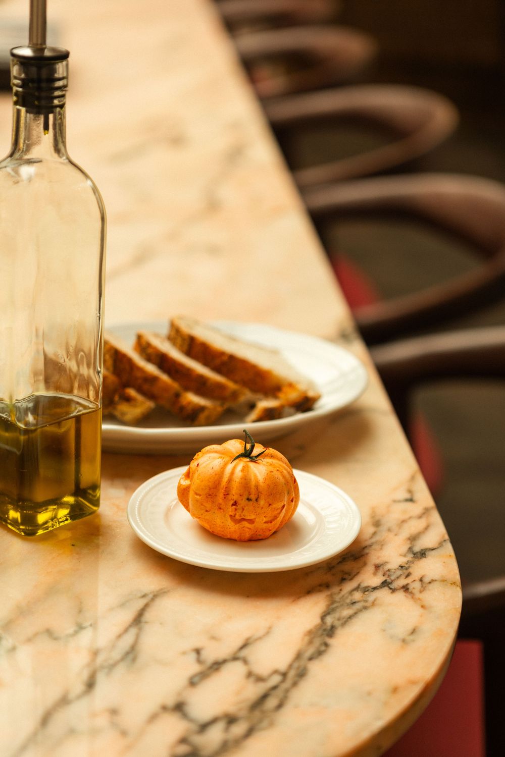 A small decorative pumpkin on a white plate next to an olive oil bottle and bread slices on a marble table.