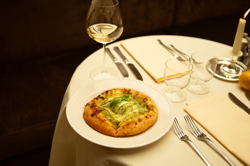 A gourmet flatbread pizza with fresh greens sits on a white plate alongside a glass of white wine on an elegant table.
