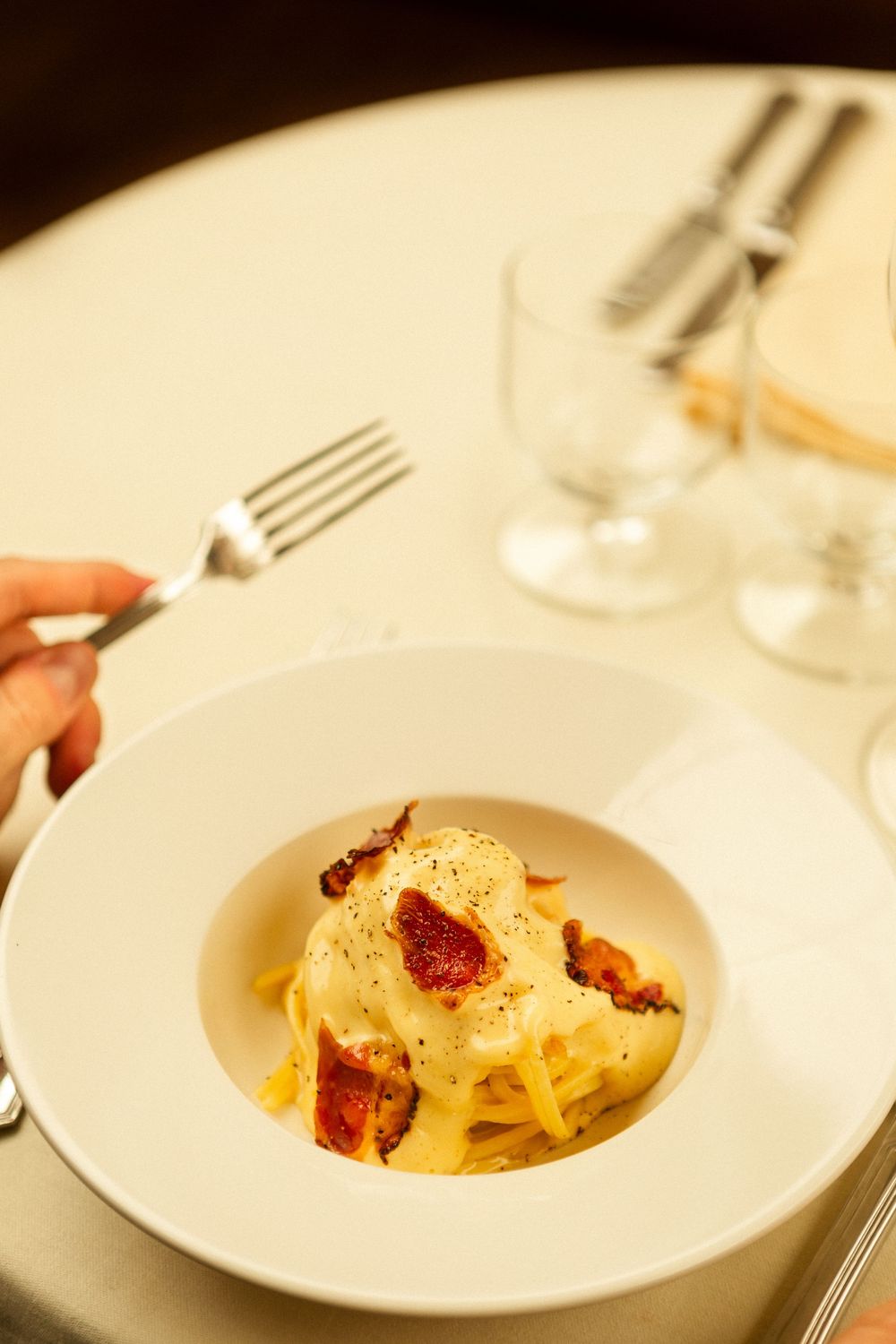 A plate of pasta carbonara garnished with bacon and black pepper on a white tablecloth.