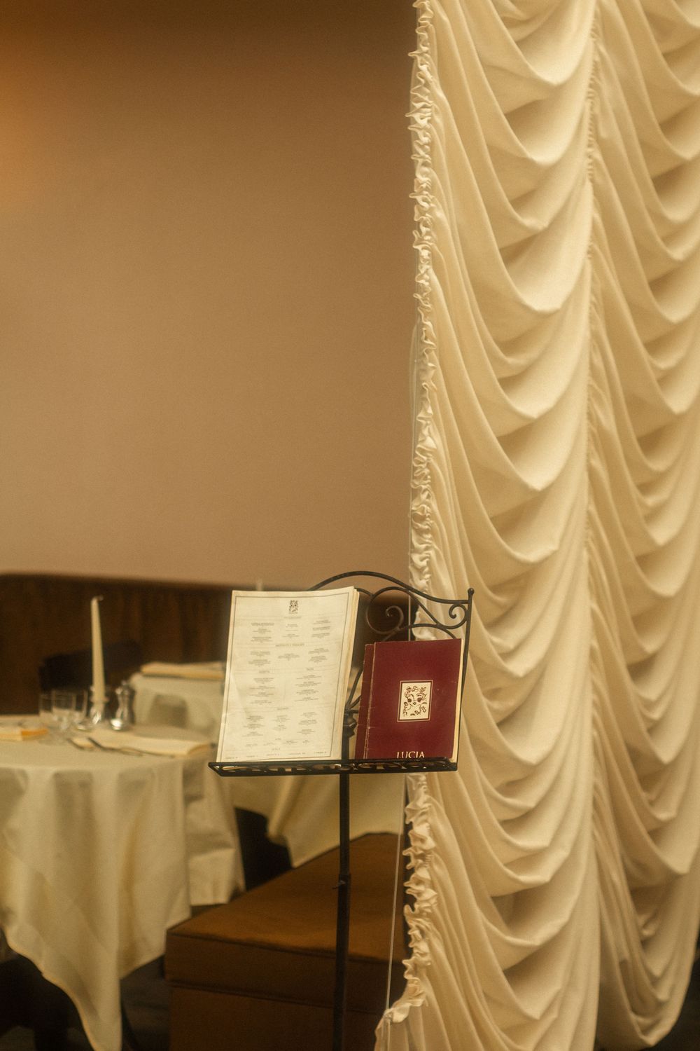 Four elegant tables with white tablecloths and music stands are arranged in an upscale dining room with draped curtains.