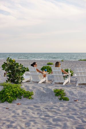 White adirondack chairs arranged on beach sand with ocean view and coastal plants.