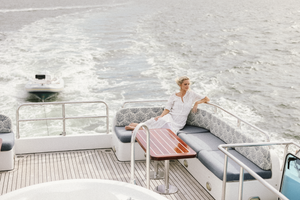 Person in white dress relaxing on deck of moving yacht with wake visible in ocean.