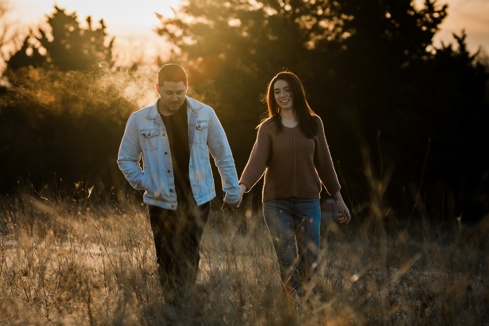 Couple holding hands while walking through a golden field at sunset with evergreen trees in background.
