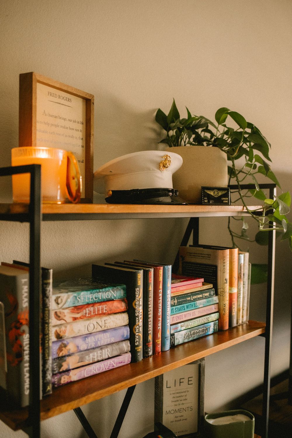 Wooden bookshelf displaying colorful novels, a white US Marine dress cover hat, candle, and green plants.