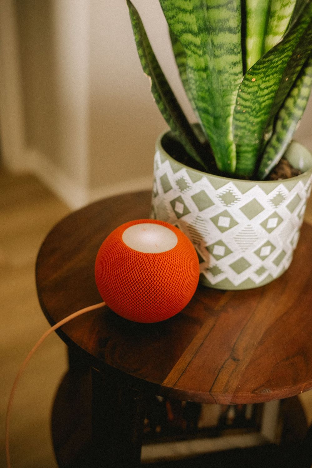 Orange Apple HomePod mini speaker on a wooden side table next to a snake plant in a geometric pot.