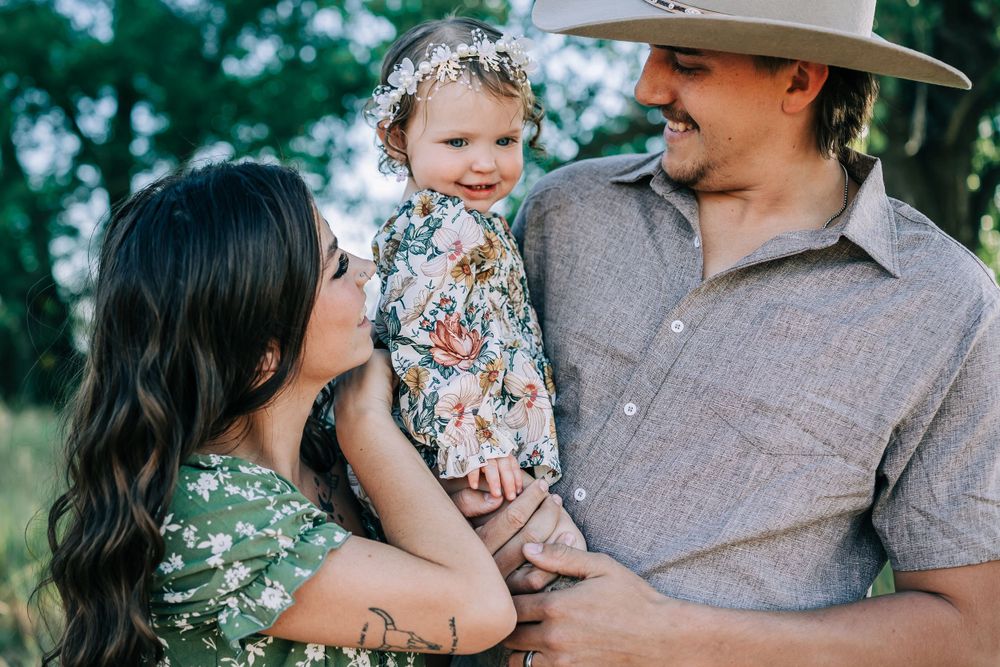 A family poses together outdoors with a small child wearing a floral dress and floral crown.