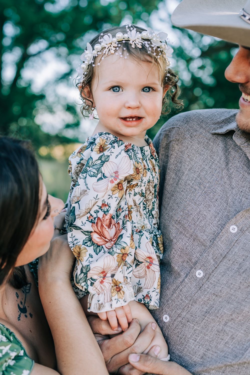 A toddler in a floral dress and baby's breath crown smiles while being held by parents outdoors.