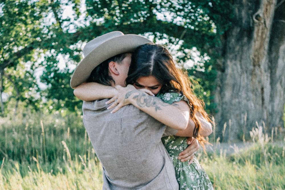 A couple embraces lovingly in a sunlit field with tall grass and trees in the background.