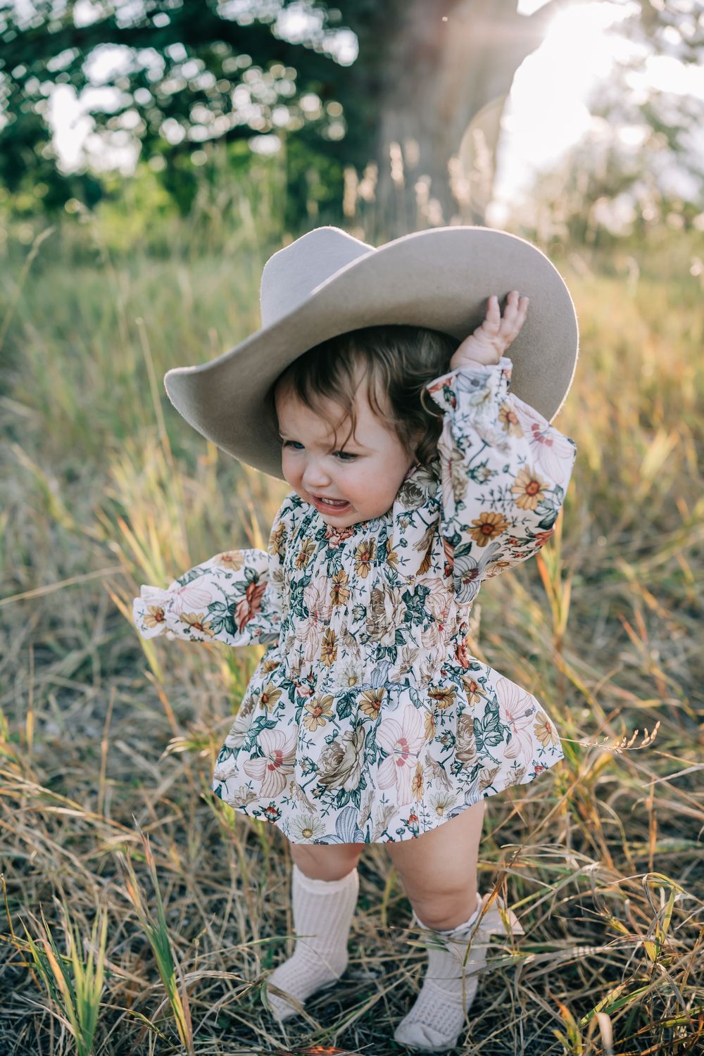 A toddler in a floral dress and cowboy hat dances joyfully in a sunny meadow.
