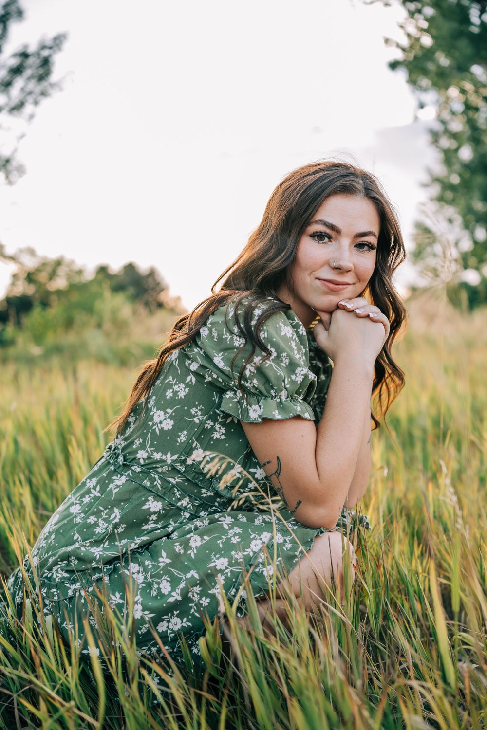 Woman in green floral dress poses gracefully in tall summer grass during golden hour.