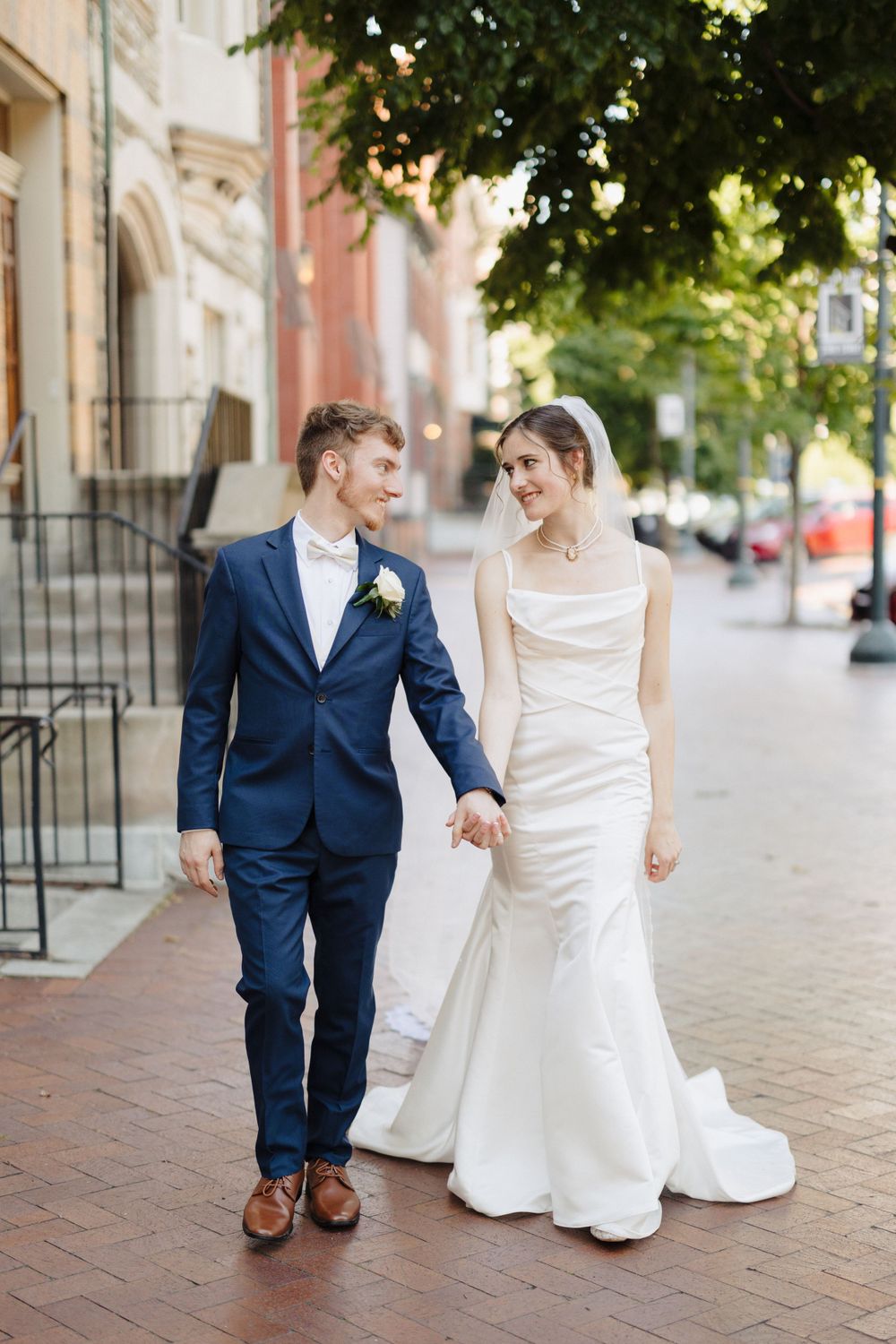 A couple in wedding attire walking together on a brick sidewalk with historic buildings in background.