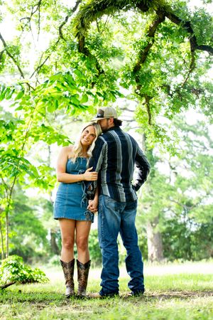 A couple in casual western attire embraces under green trees in a romantic outdoor setting.