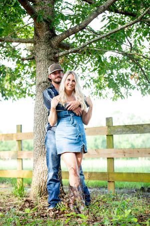 A couple in western attire poses together against a large tree and wooden fence.
