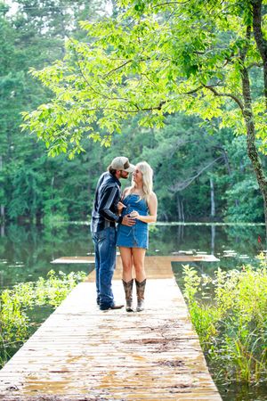 A couple stands close together on a wooden dock surrounded by lush summer scenery.