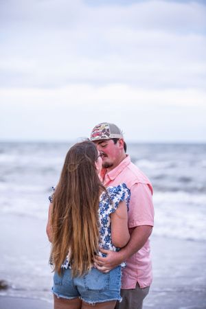 A romantic couple embraces on a cloudy beach with waves crashing in the background.