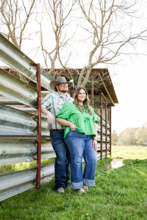 Two people lean against a metal fence near a rustic barn structure.