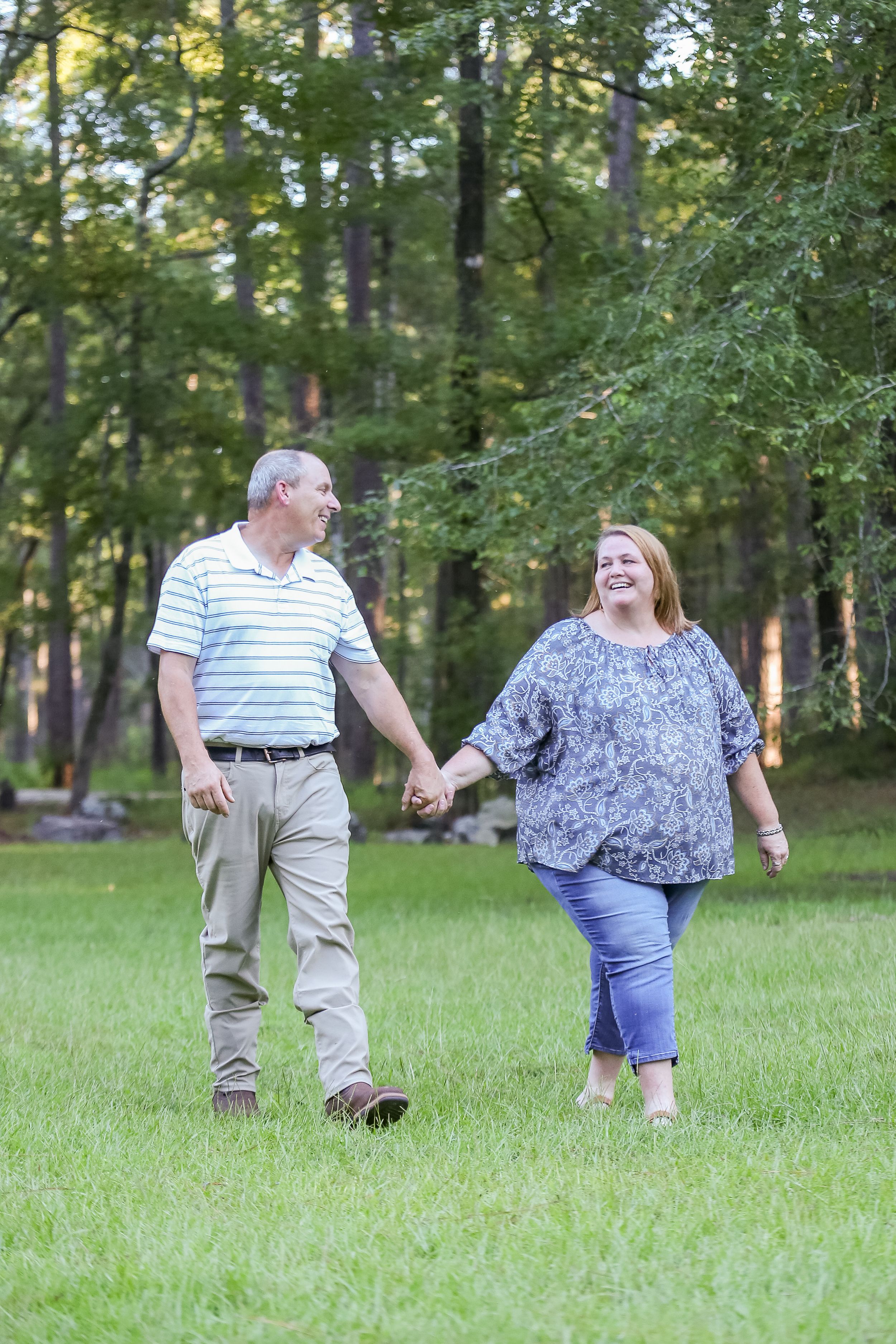A couple walks hand in hand through a sunlit grassy area surrounded by trees.