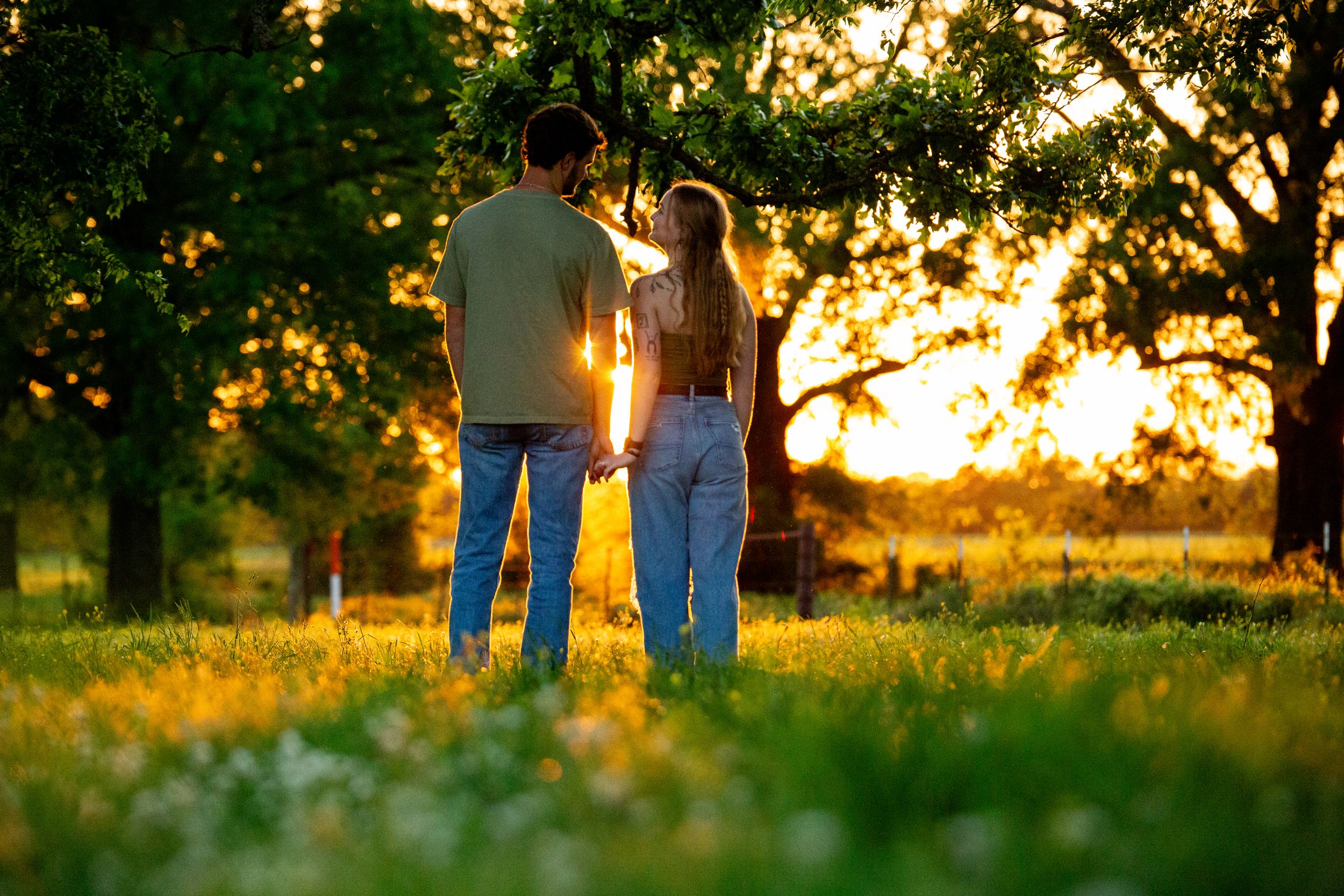 Couple walking through a grassy field at sunset with trees in the background.