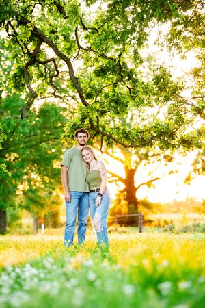 Couple standing together in a lush green field with sunlit trees behind them.