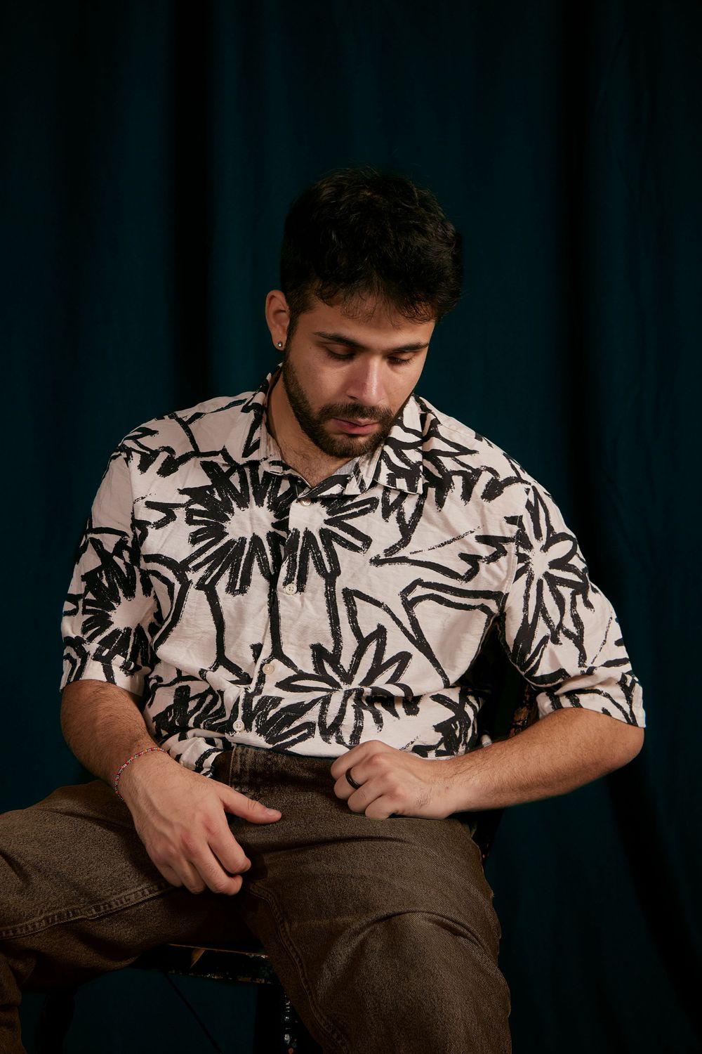 Studio Portrait of a man sitting on a stool, looking towards the ground with his hands in his lap.