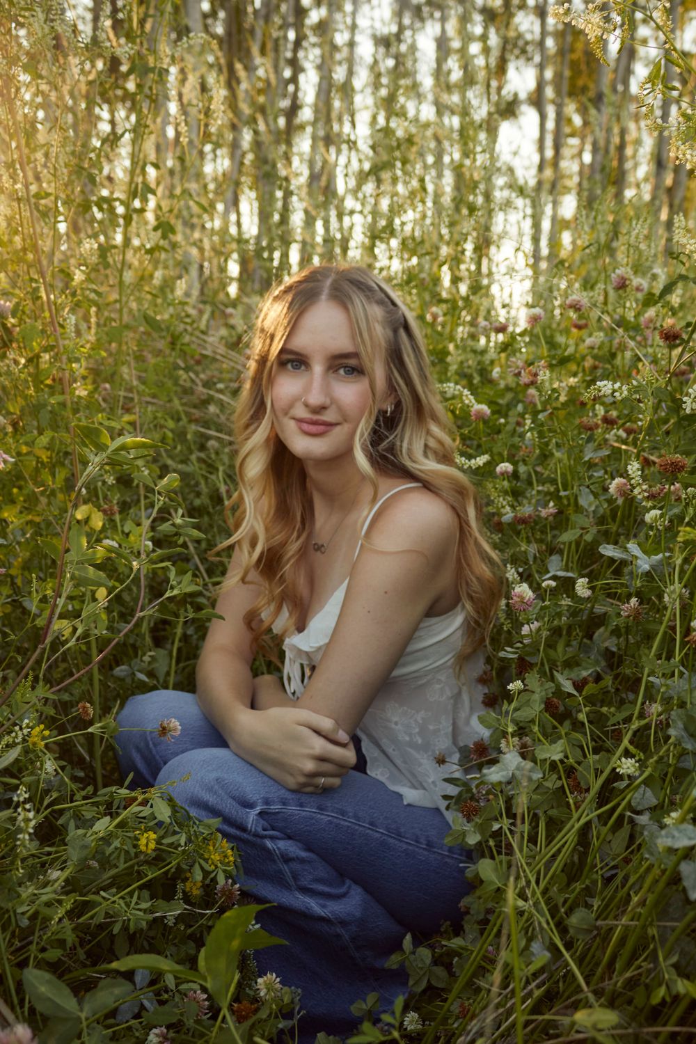 Young woman sitting in a field surrounded by flowers. She has curled hair and is wearing jeans and a tank top