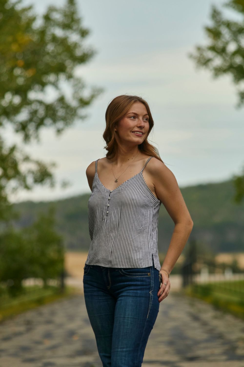 Young woman standing in the middle of a road with trees. Wearing a tank top and jeans.