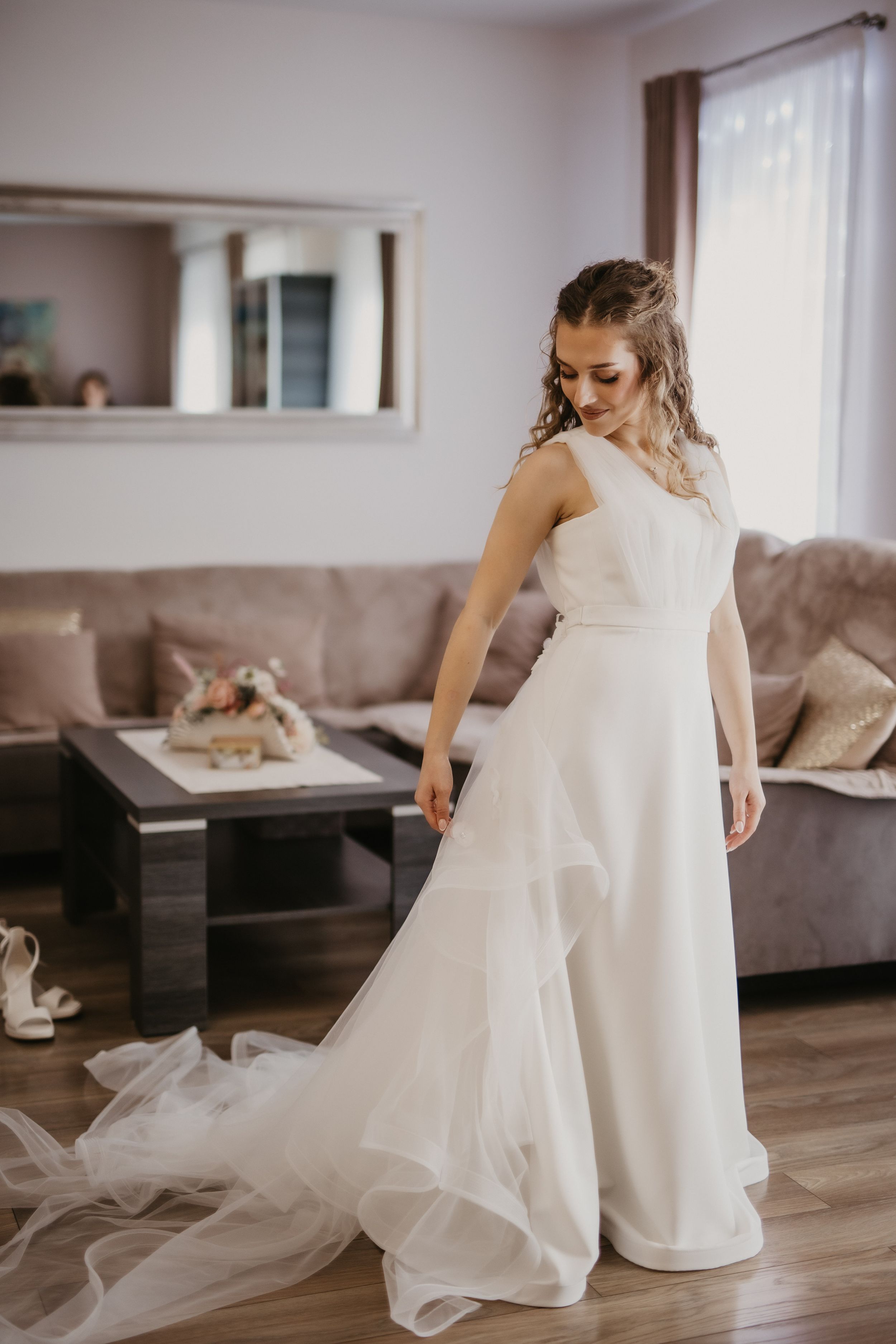 A bride in an elegant white wedding gown standing in a modern living room with natural light.