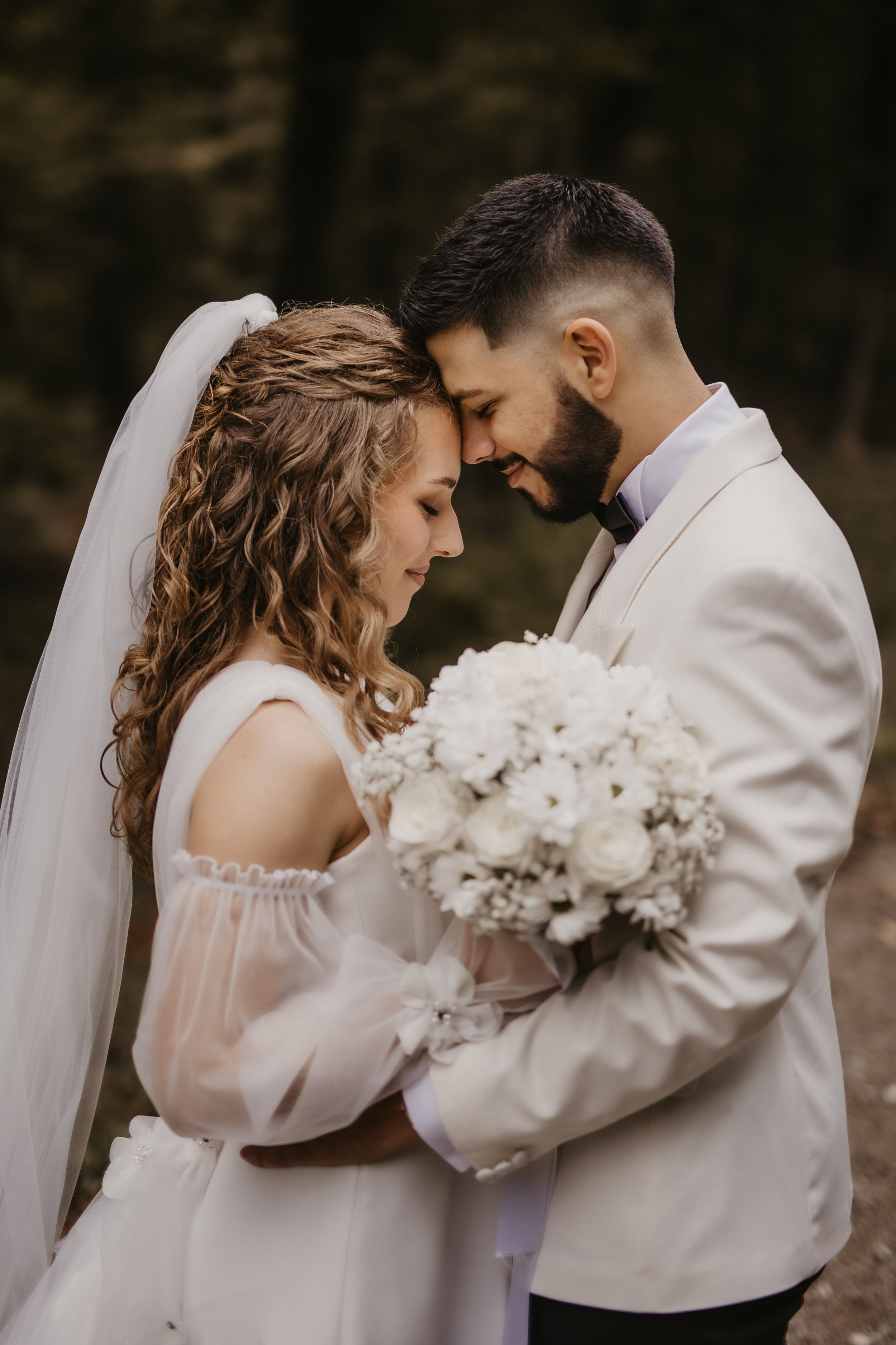 Intimate wedding couple embraces while holding white bouquet in moody forest setting.