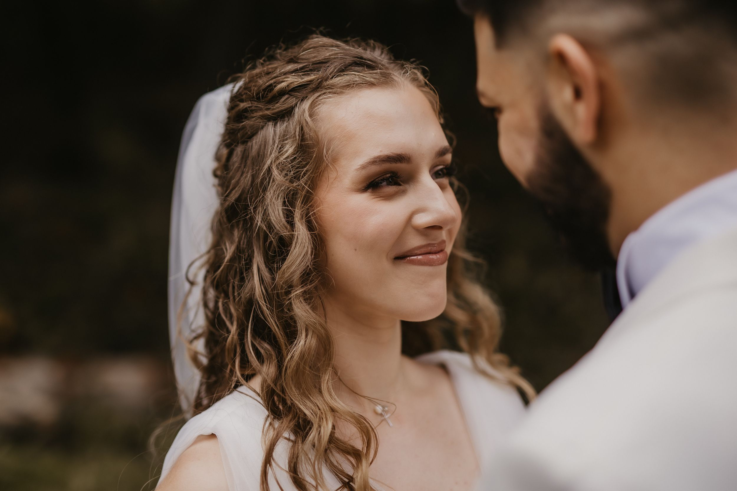 Close up of bride with curly hair smiling at groom during outdoor wedding.