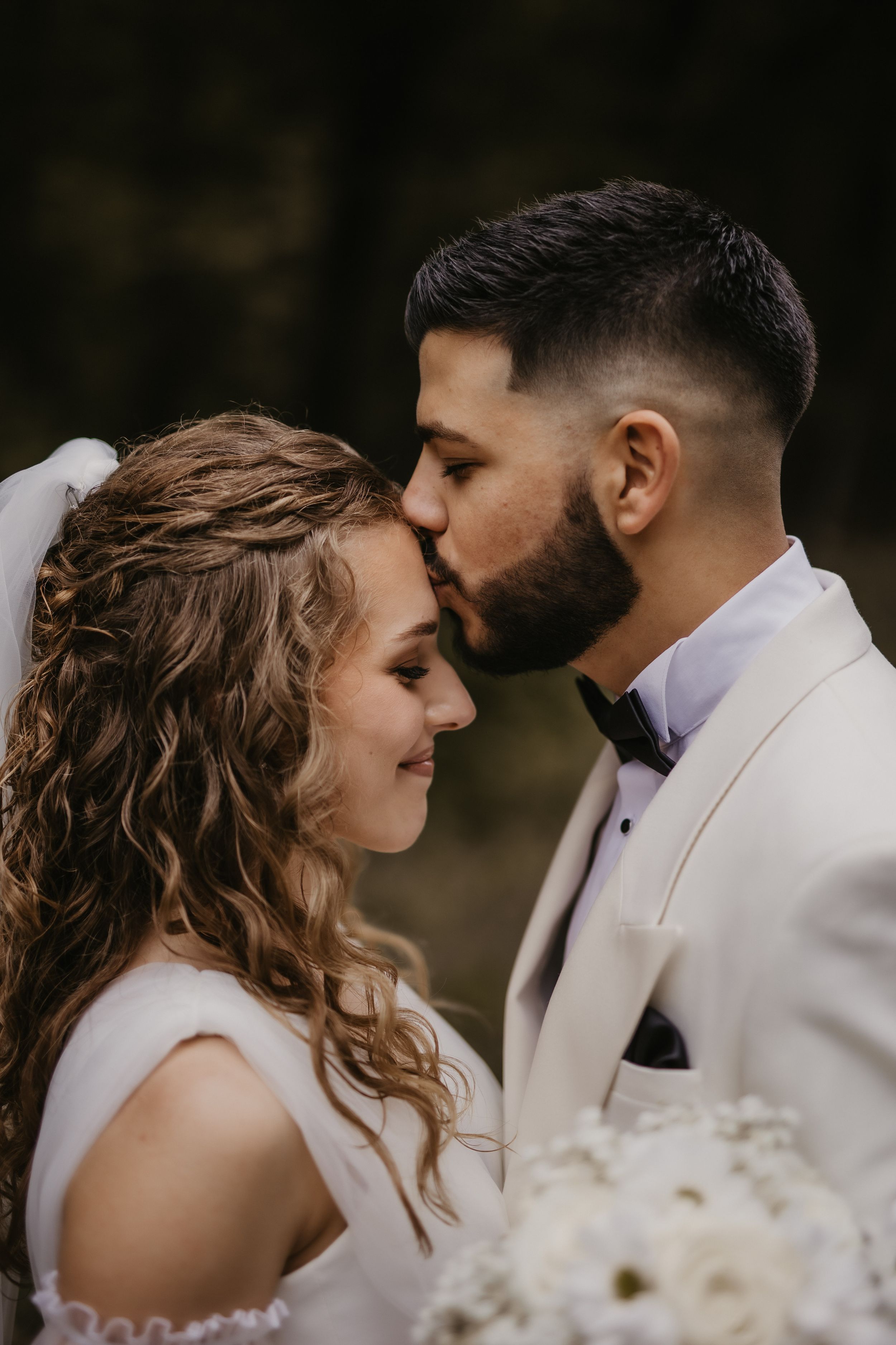 Romantic wedding moment with groom kissing bride's forehead during forest photoshoot.
