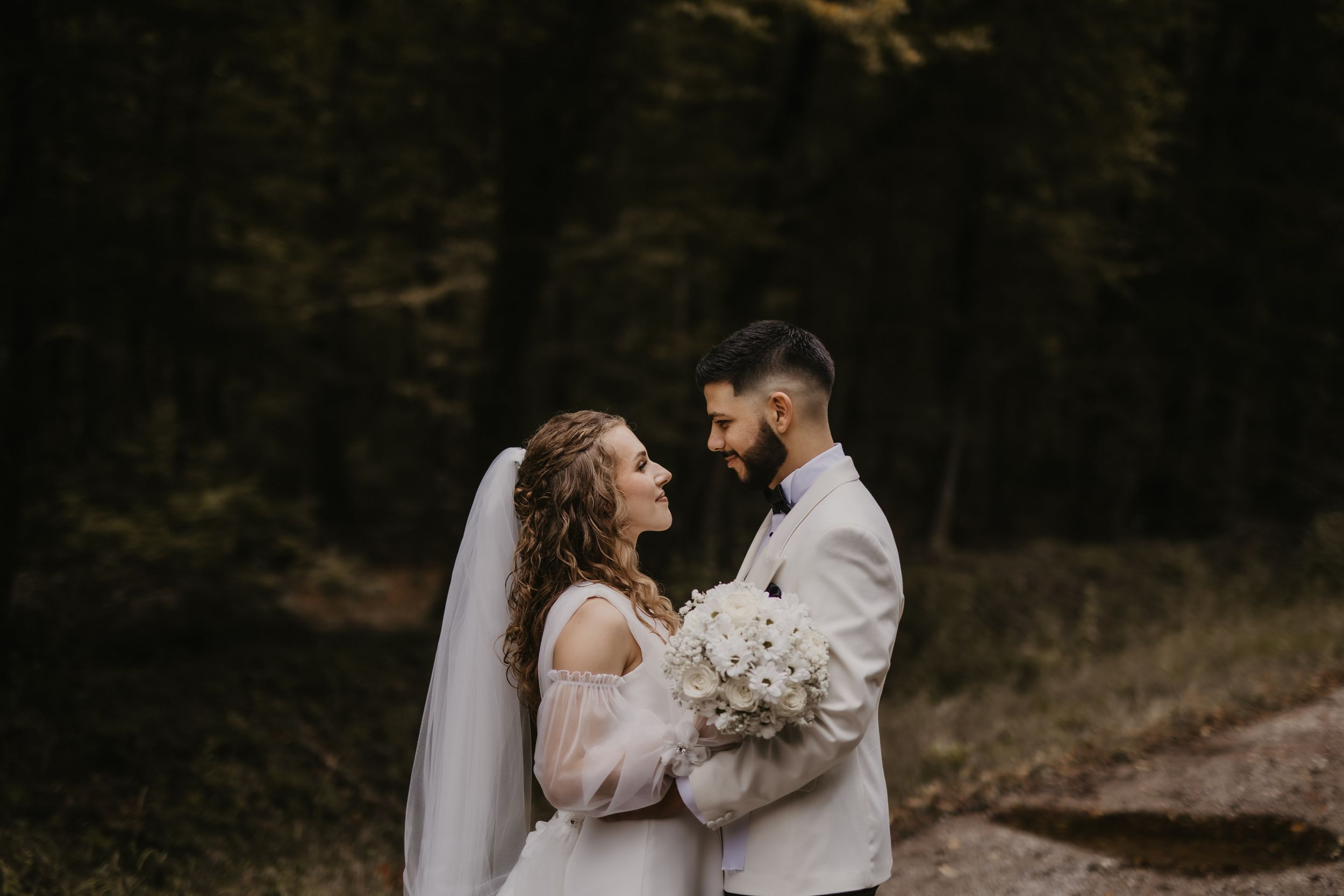 Wedding couple shares tender moment in dark wooded setting with white floral bouquet.