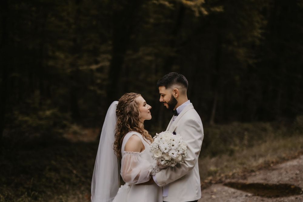 Wedding couple shares tender moment in dark wooded setting with white floral bouquet.