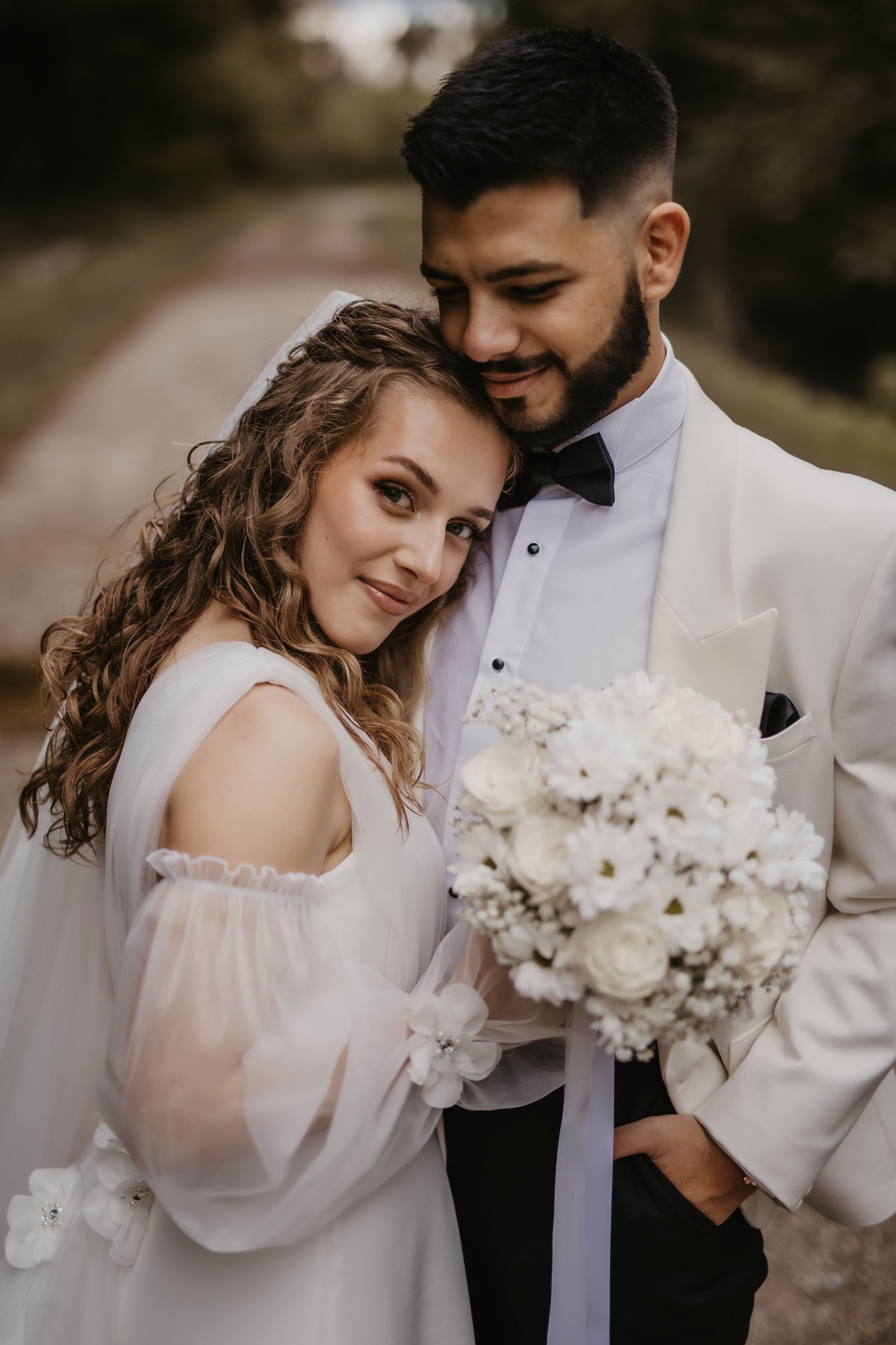 Bride in white dress holds bouquet while leaning against groom in white jacket.