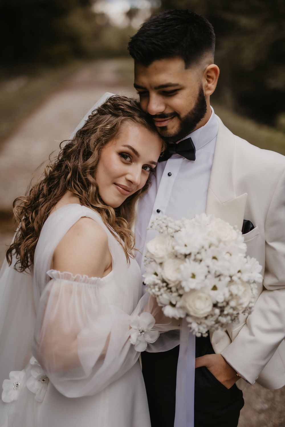 Bride in white dress holds bouquet while leaning against groom in white jacket.