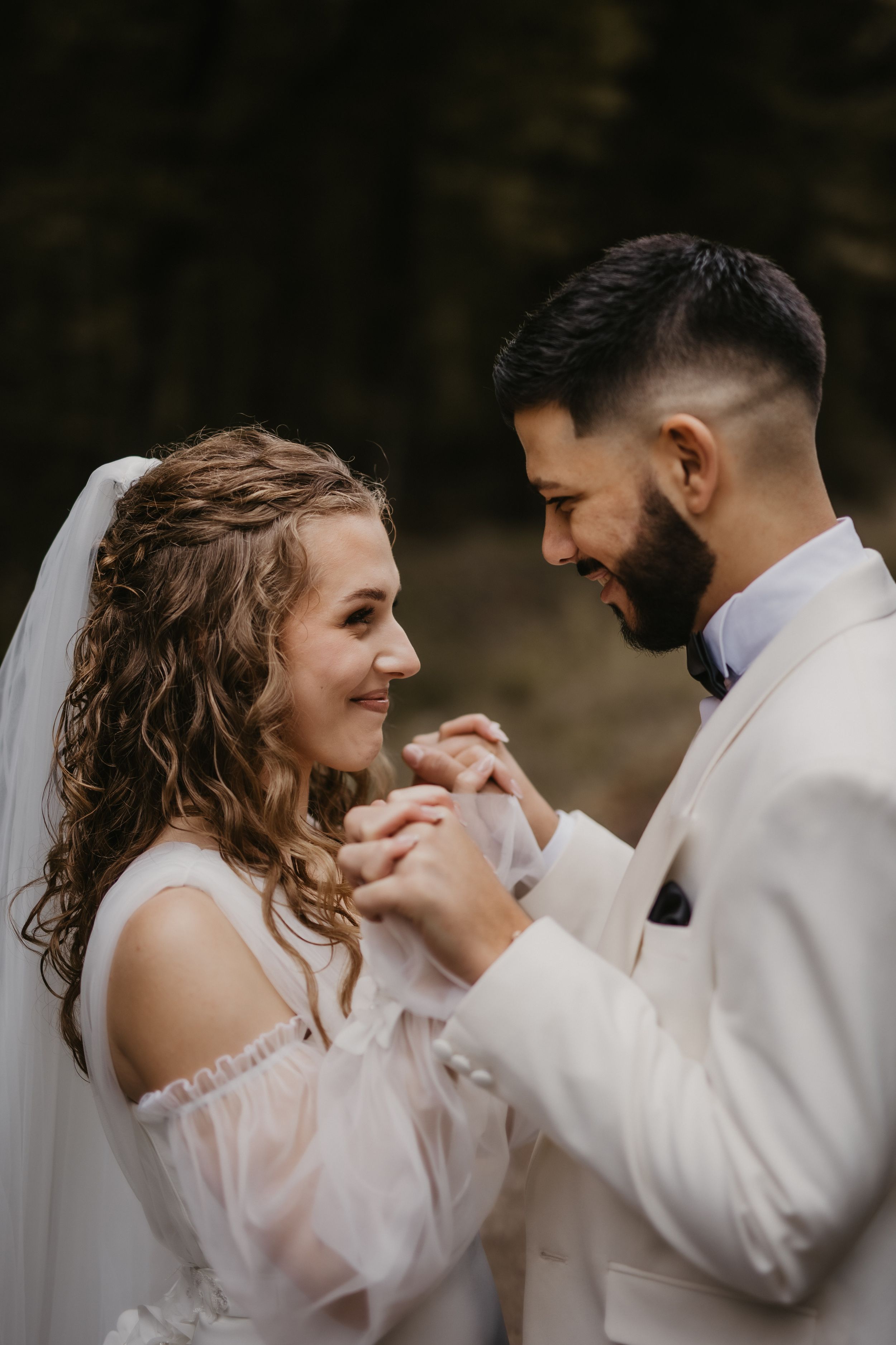 Wedding couple holds hands and smiles at each other during intimate outdoor moment.