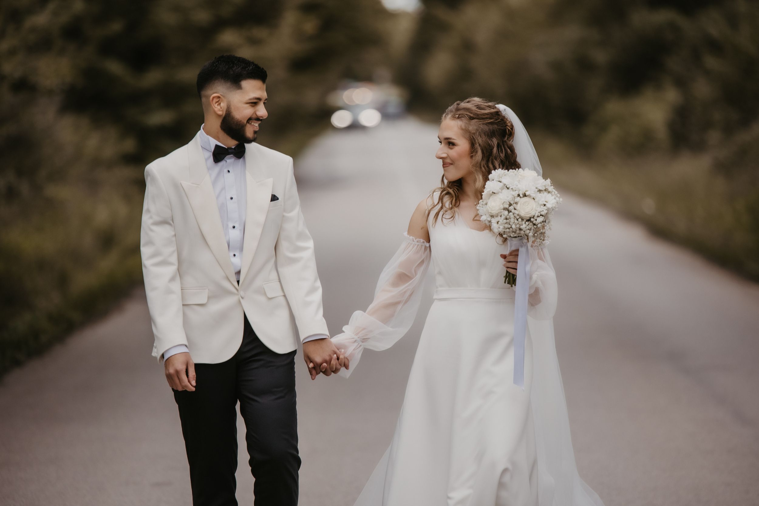 Bride and groom in white attire walk hand in hand down country road.