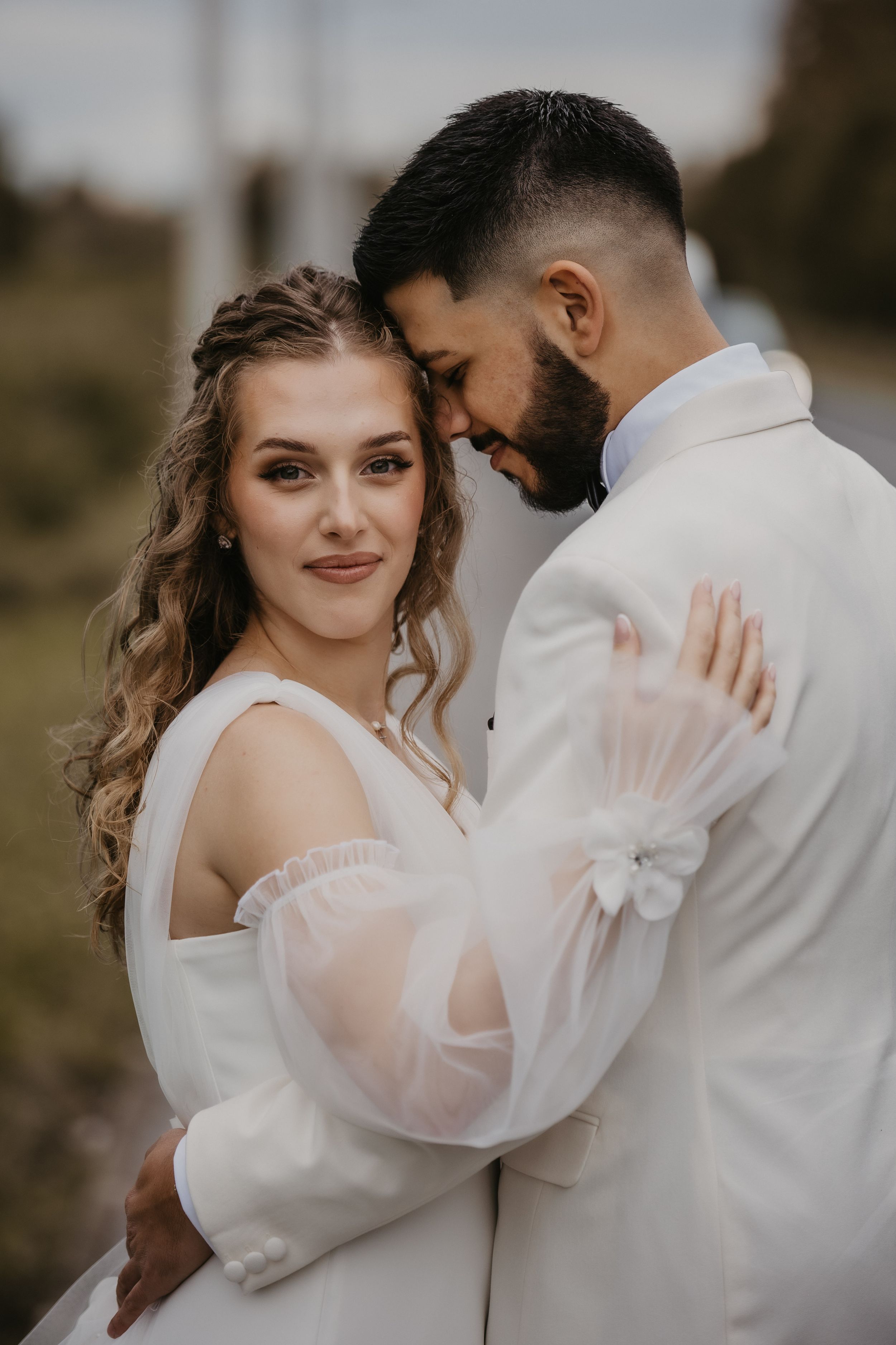 Intimate embrace between newlyweds in white wedding attire against natural backdrop.