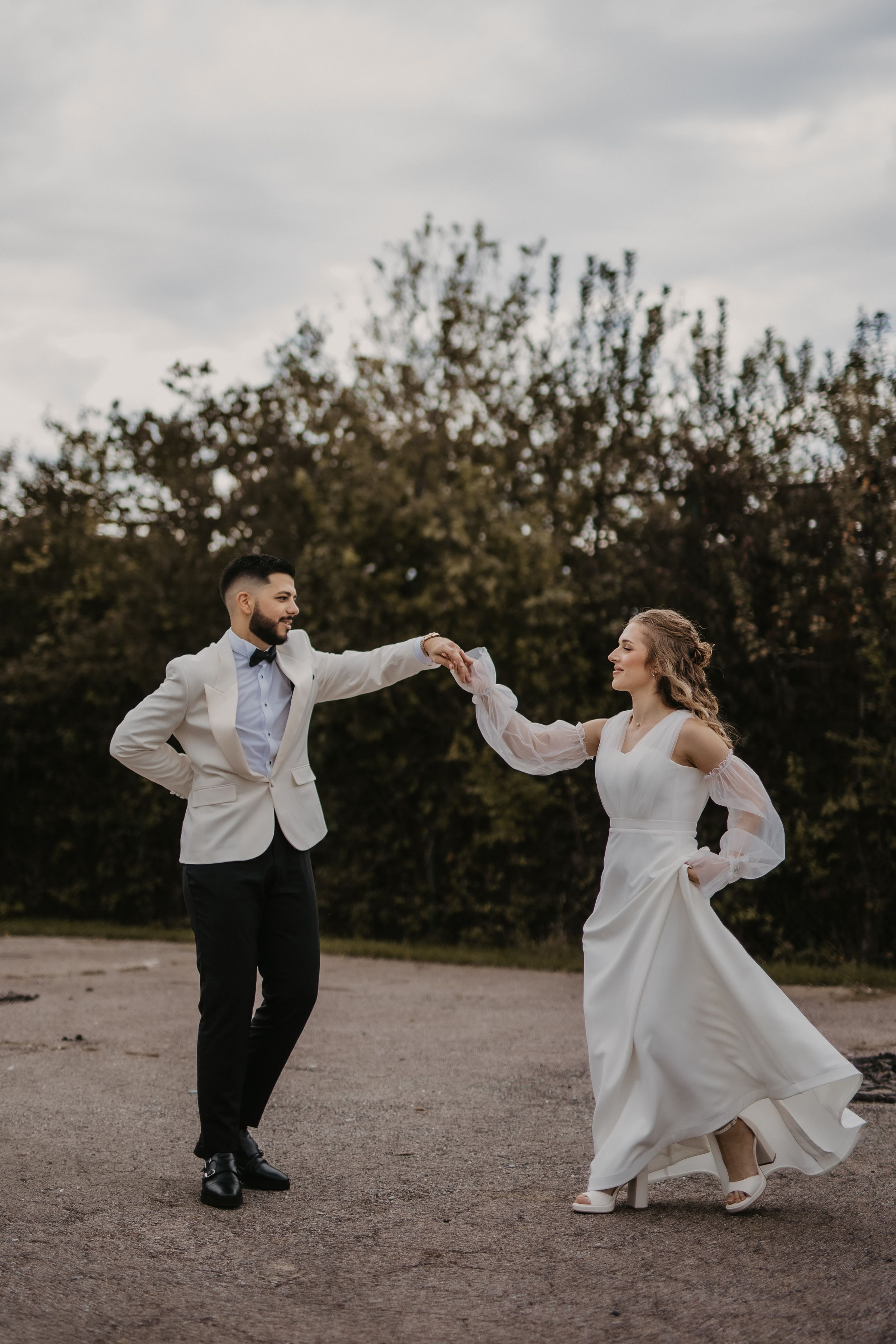 Joyful couple in wedding attire dancing on dirt path with trees in background.
