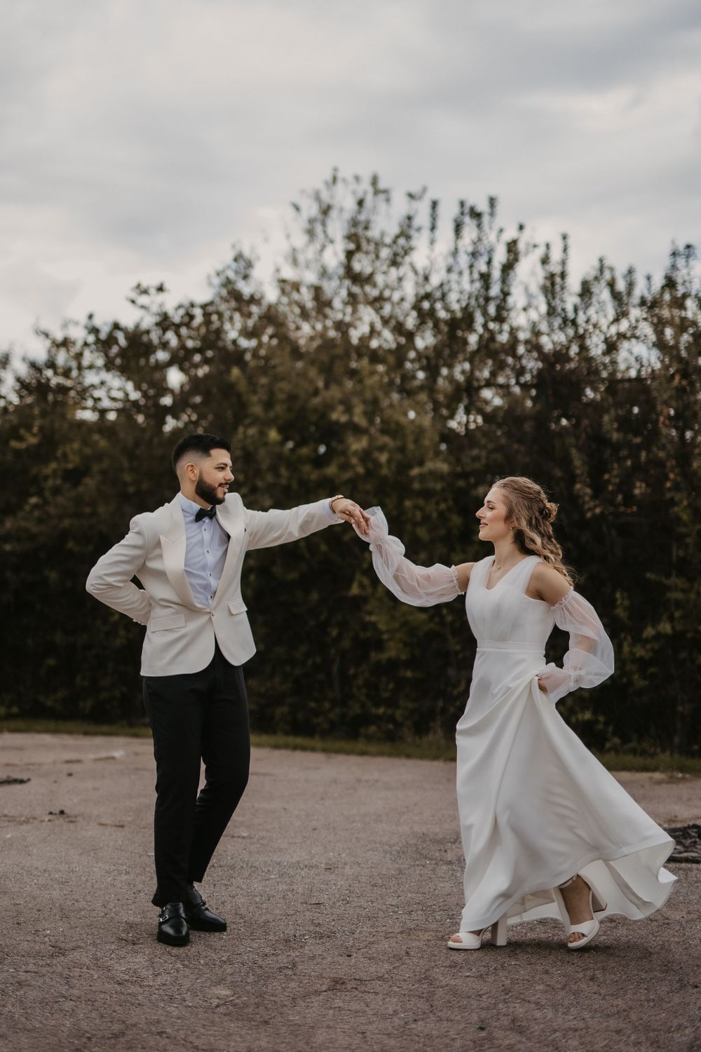 Joyful couple in wedding attire dancing on dirt path with trees in background.