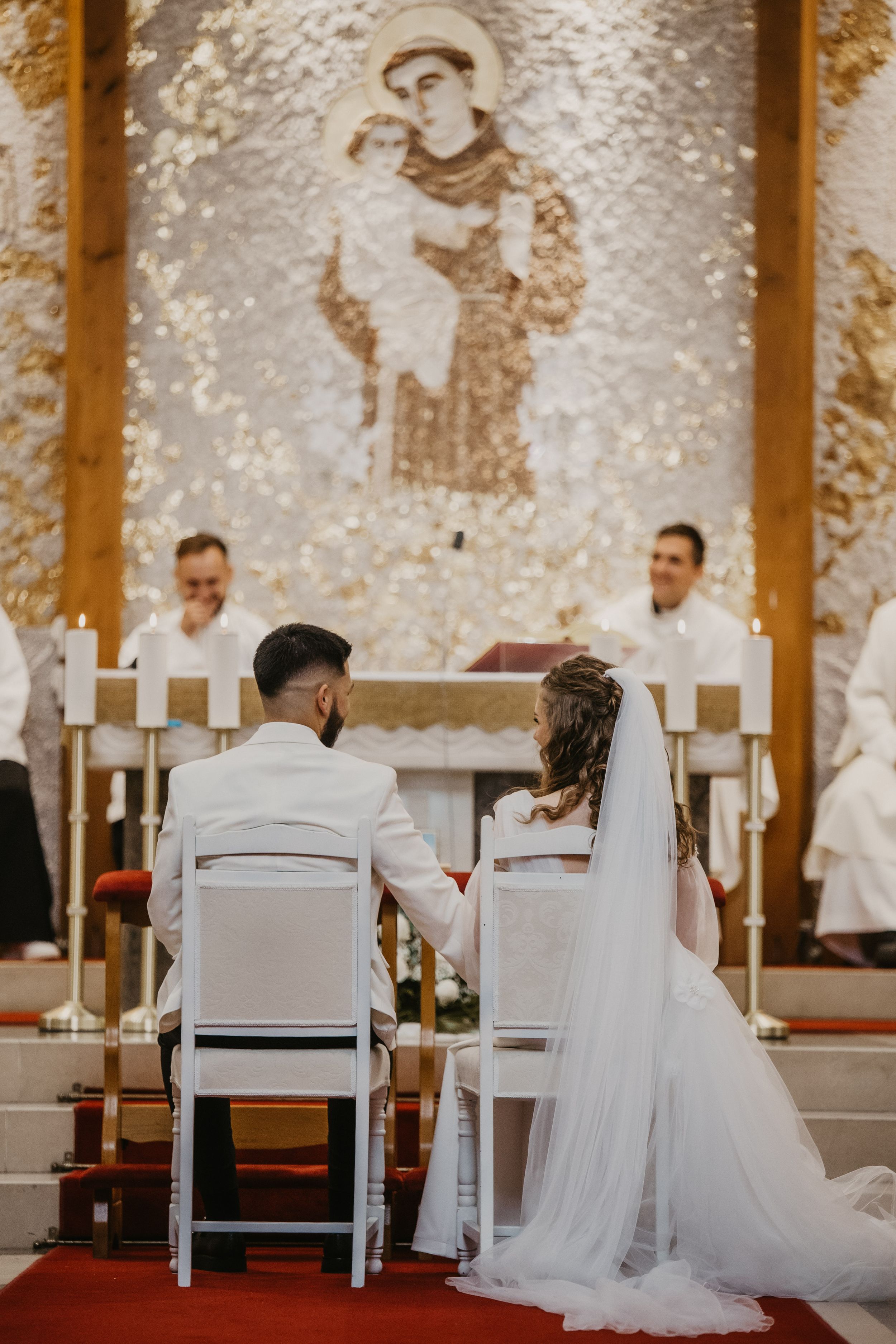 A couple sits in white chairs during their wedding ceremony with a religious mural in the background.