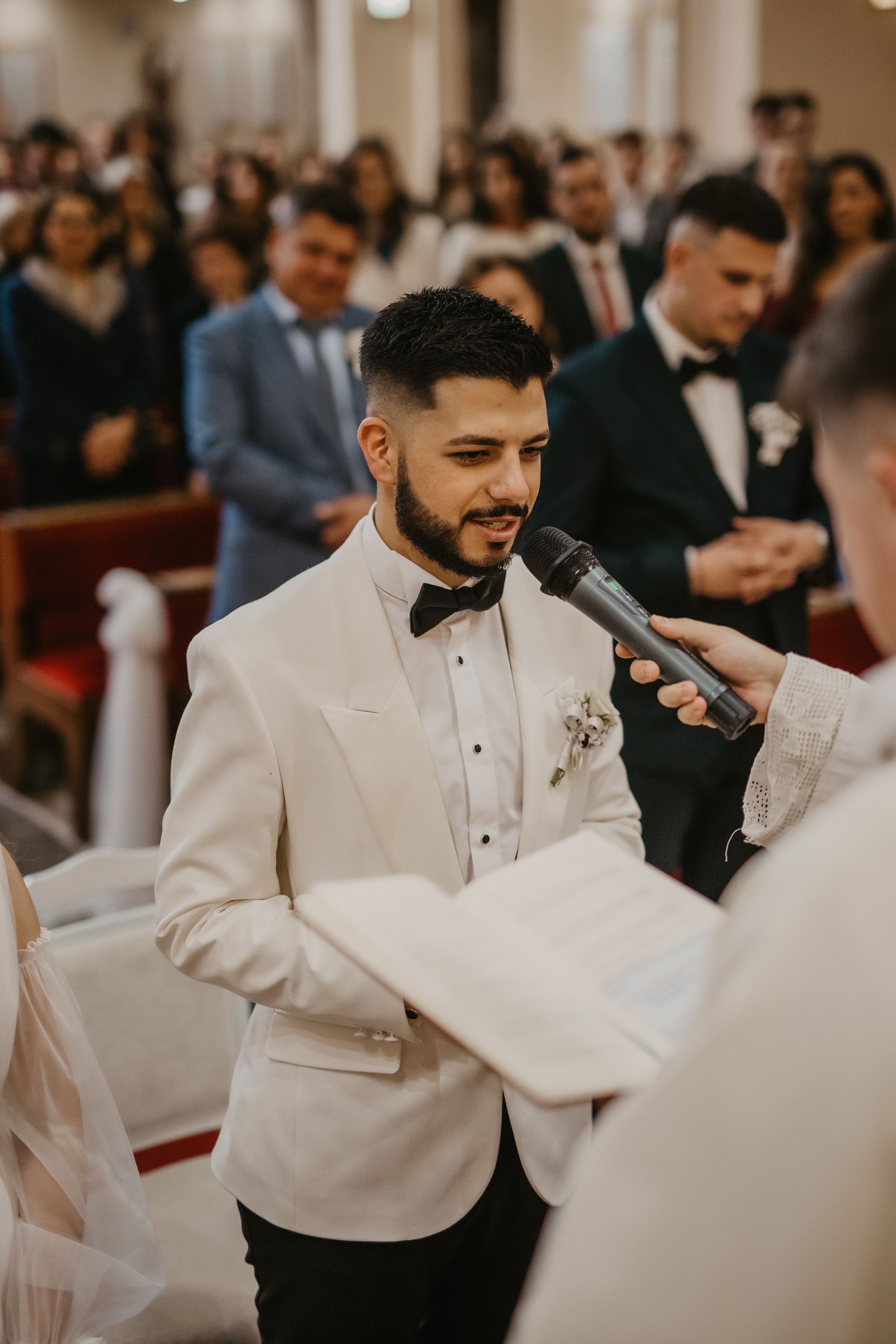 A groom in a white tuxedo jacket reads vows during a church wedding ceremony.