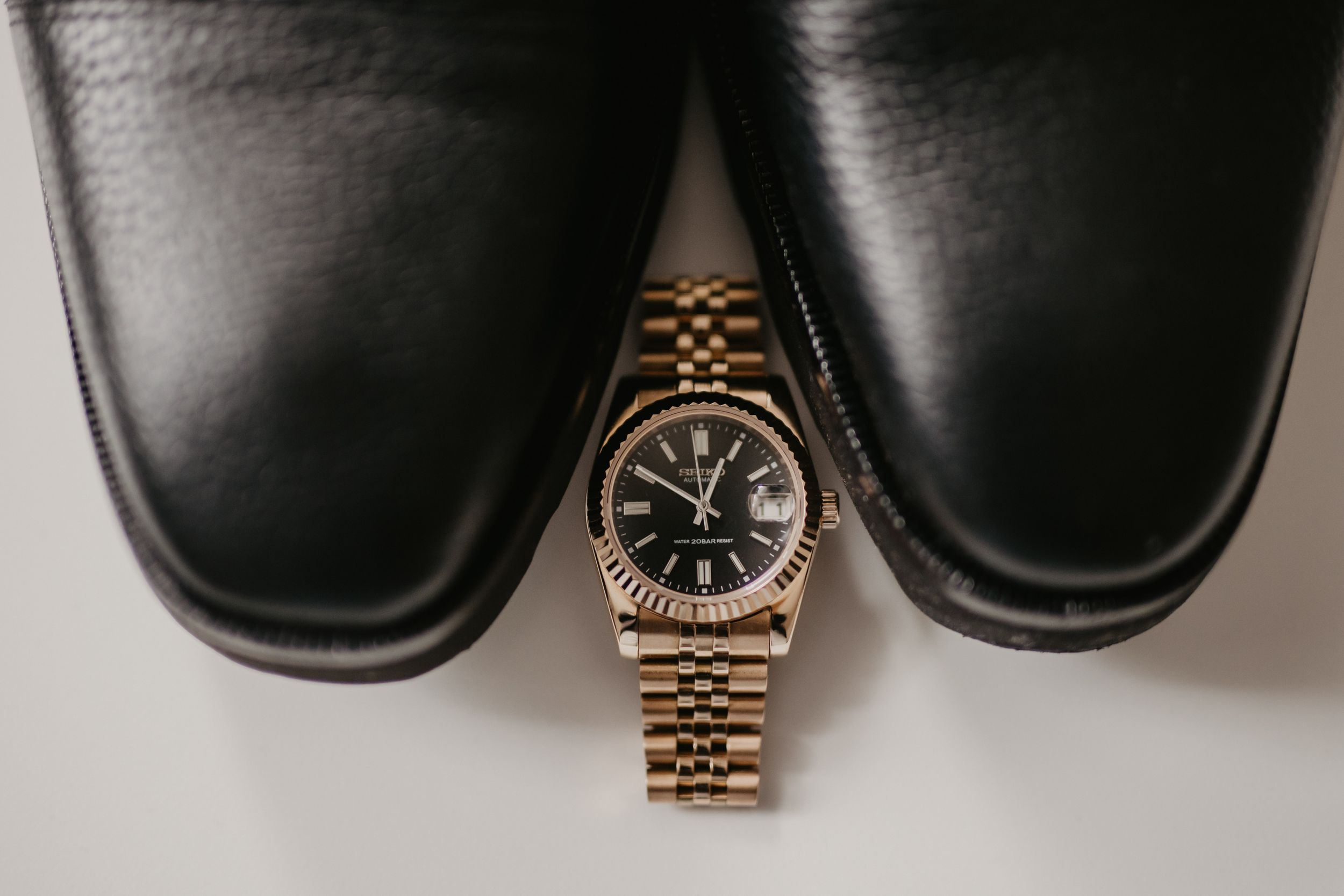 A luxury gold watch placed between formal black leather dress shoes on white background.