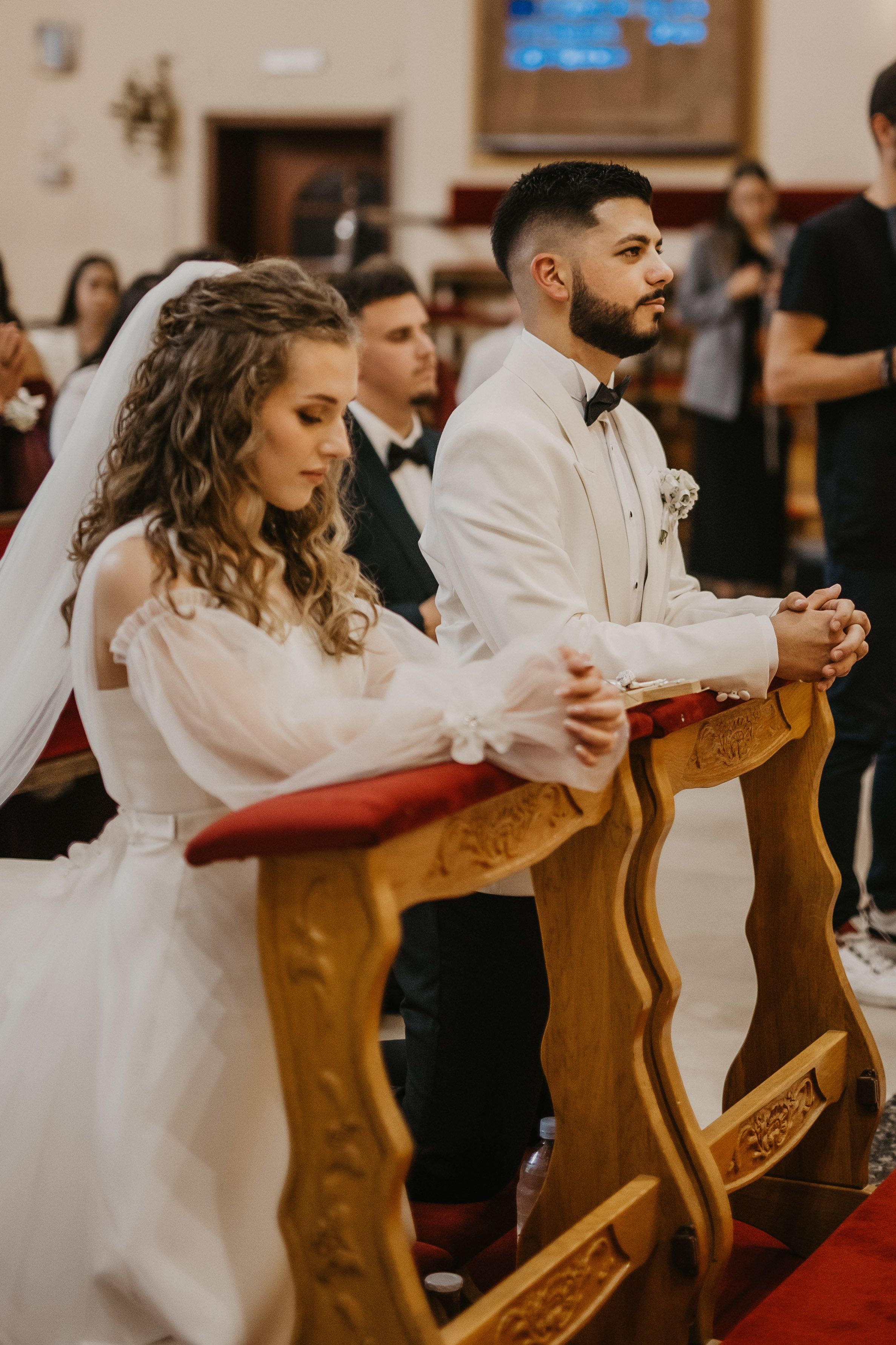A couple kneels at ornate wooden prayer benches during their church wedding ceremony.