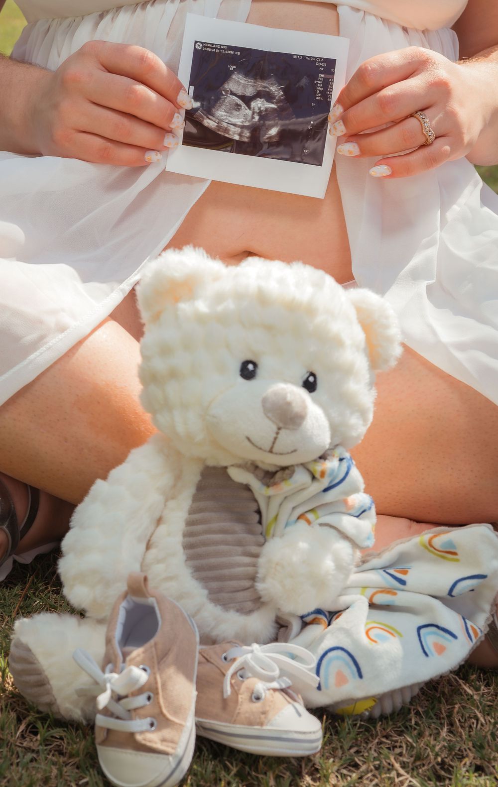 Hand holding ultrasound photo with white teddy bear and baby shoes in foreground showing pregnancy announcement scene.